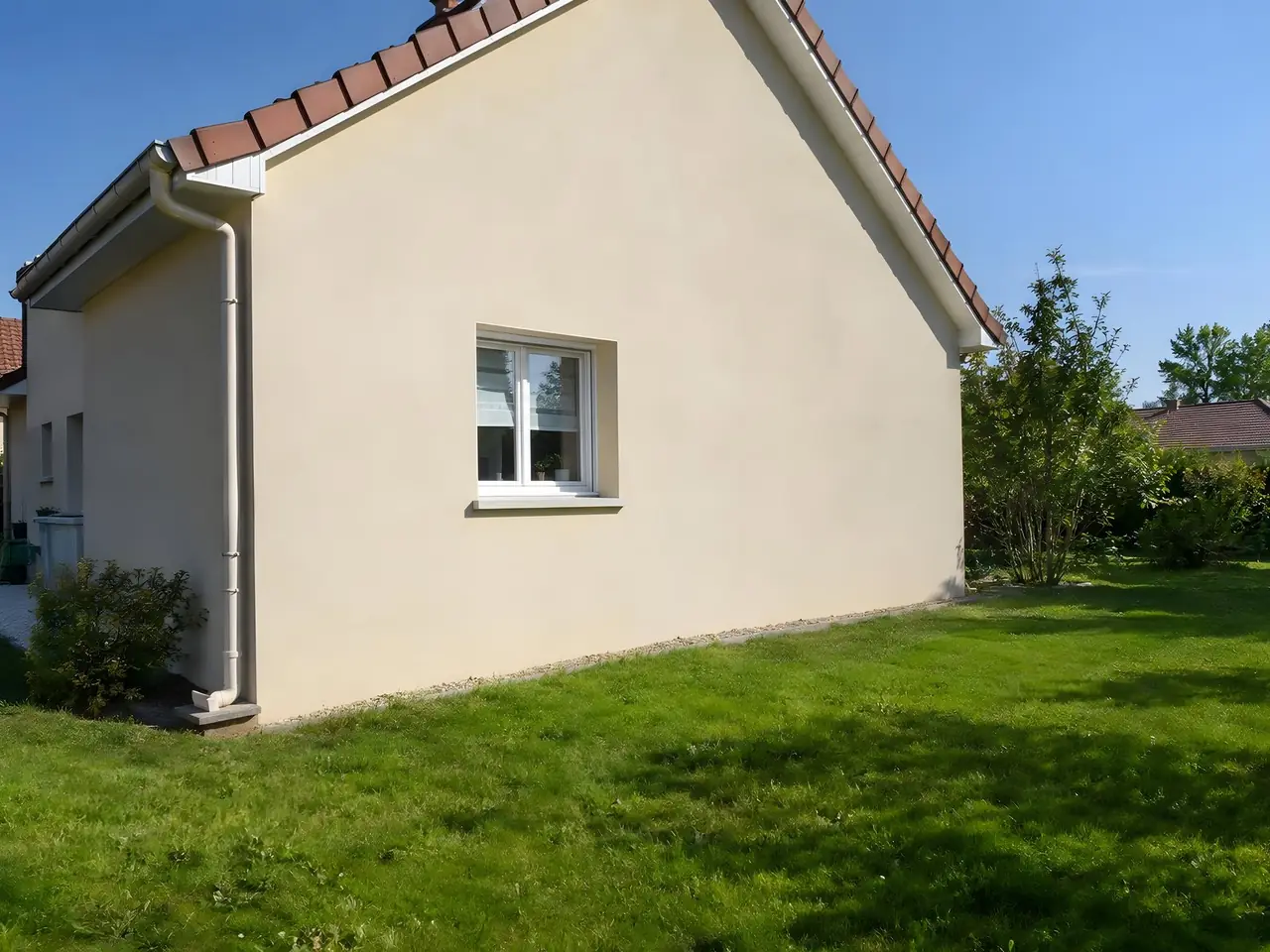 Pignon crépi beige d'un pavillon en plein soleil sous un toit de tuiles rouges, pelouse verte dégagée au pied du mur