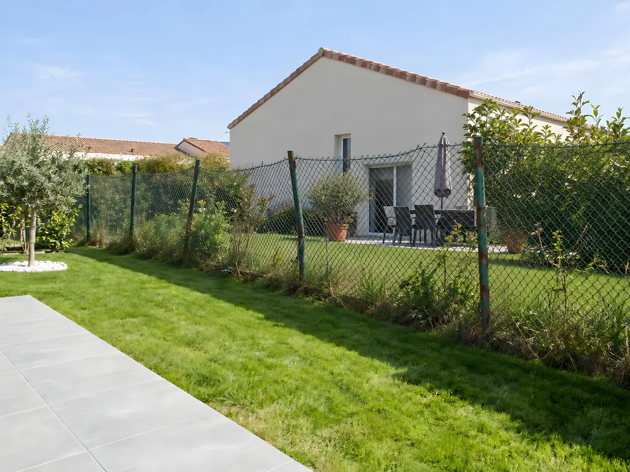 Vieux grillage souple vert affaissé sur des poteaux tordus, envahi d'herbes hautes, entre une pelouse et le jardin voisin